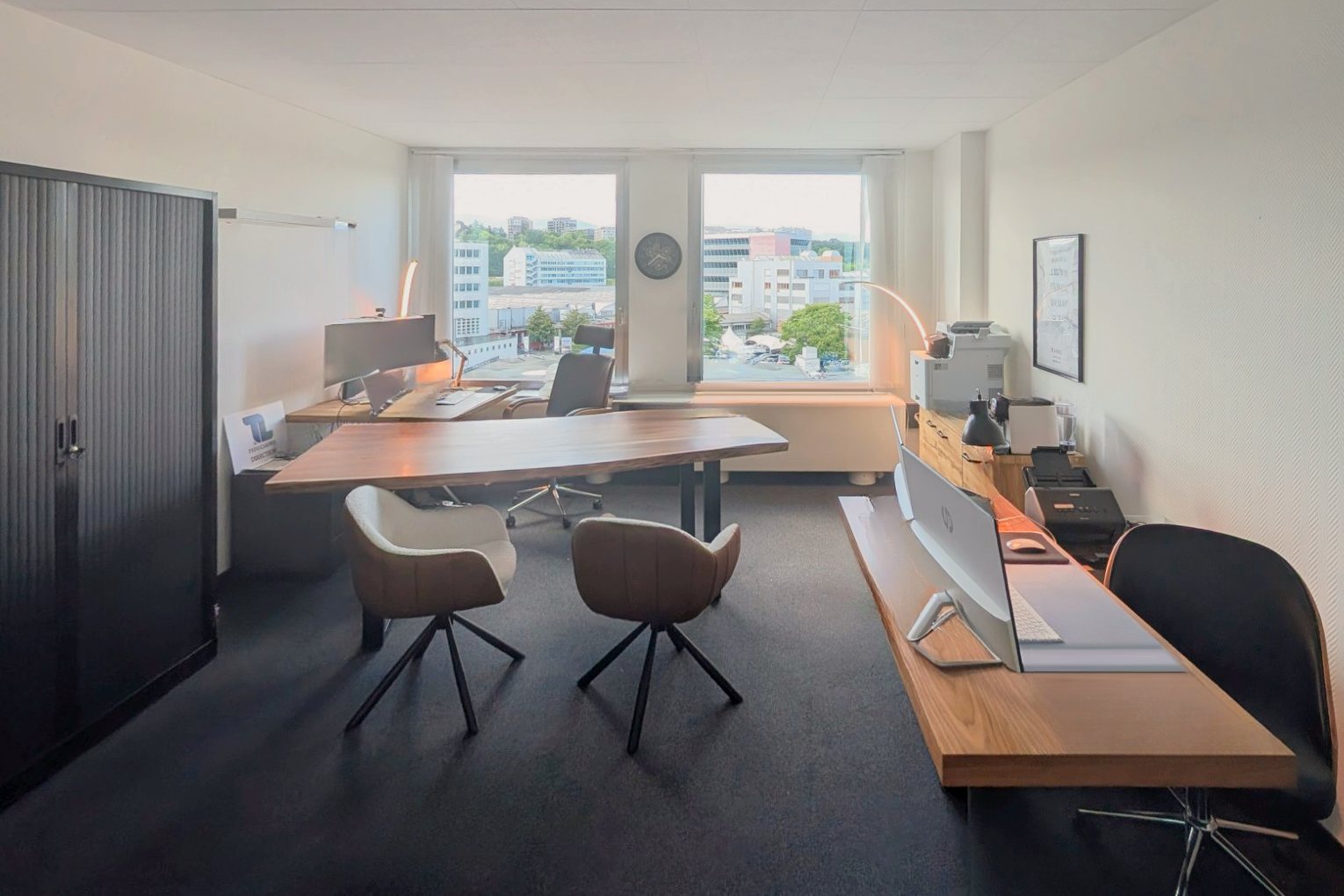 Bureau de la direction de TL Fiduciaire à Carouge : grande table en bois devant deux fenêtres qui donnent sur des immeubles, deux fauteuils visiteurs, un poste de travail avec grand écran au fond, une armoire noire à gauche et, à droite, un bureau avec ordinateur et imprimante.